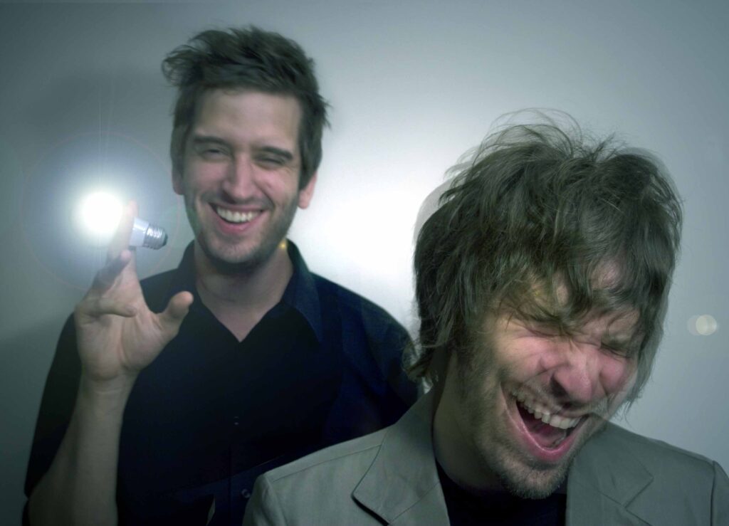 Two men laugh together; one holds a small glowing lightbulb, while the other bends forward, eyes closed and mouth open in laughter. The background is neutral and softly lit.