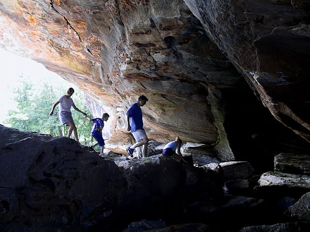 Four people explore a rocky cave, with light shining in from the left. They are climbing over large rocks with the cave ceiling arching above them. Trees are visible outside the cave entrance.