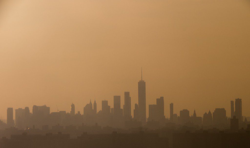 Silhouetted city skyline at sunset, with buildings and skyscrapers visible through a hazy, orange-tinted sky.