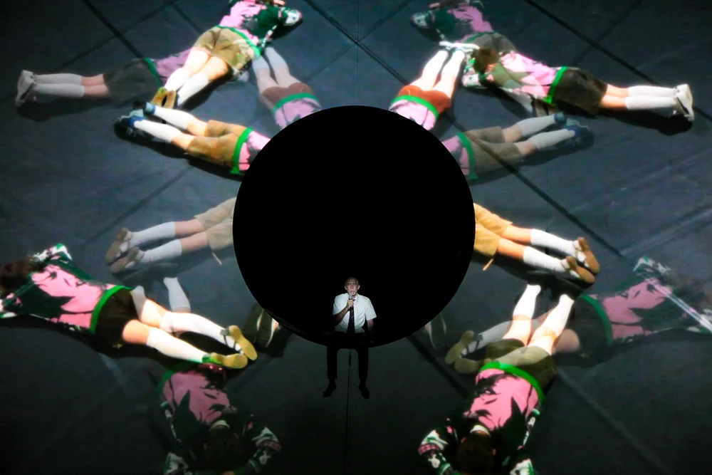 A person stands in front of a dark circle on stage, with performers in matching outfits lying in a circular pattern and their reflections visible on the shiny floor.
