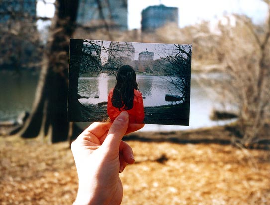 A hand holds a photo of a girl with long hair in a red coat, aligning it with the same outdoor scene in the background, blending past and present.
