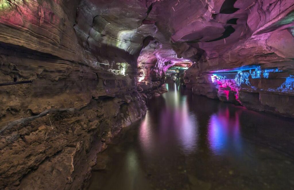 A rocky underground cave with a calm stream running through it, illuminated by colorful purple, pink, and blue lights reflecting on the water and cave walls.