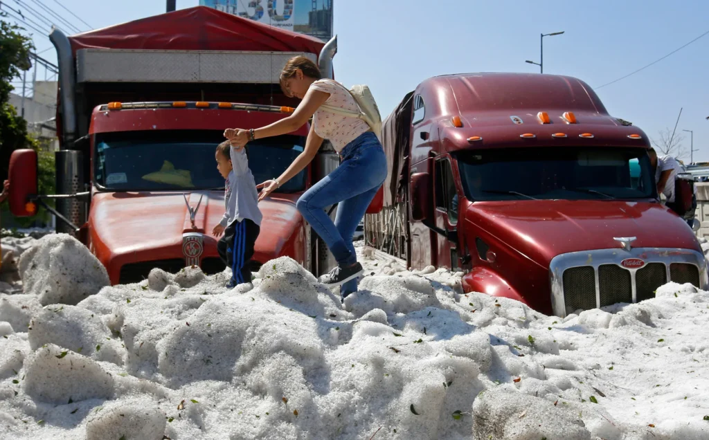 A woman helps a small child stand atop a mound of hail in front of parked red trucks on a city street after a heavy hailstorm.