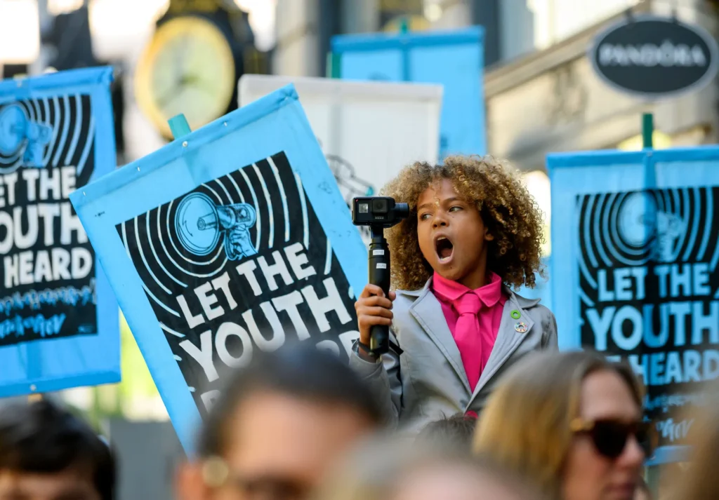 A child in a suit, holding a camera and shouting, stands among people at a rally with blue signs reading “Let the Youth Be Heard.” The scene is lively, with a focus on youth activism.