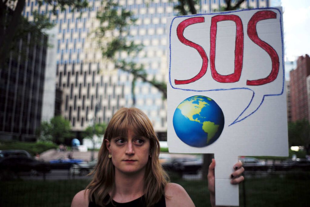 A person stands outside holding a sign with a drawing of Earth and a speech bubble containing “SOS” in red letters, with office buildings in the background.