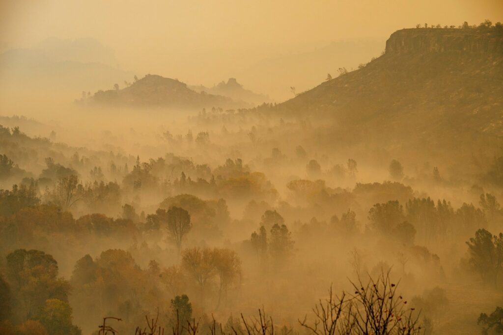 Hazy landscape with rolling hills and scattered trees shrouded in thick smoke or fog, under a yellow-orange sky, creating a moody, atmospheric scene.