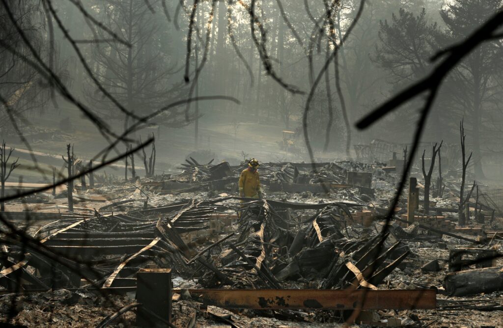 A firefighter in yellow gear stands among the charred remains of buildings, surrounded by ash, smoke, and burned trees after a wildfire.