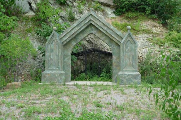 Stone archway with pointed roof stands in front of a dark, gated cave entrance surrounded by green foliage and rocky terrain. Sparse grass and small plants cover the ground.