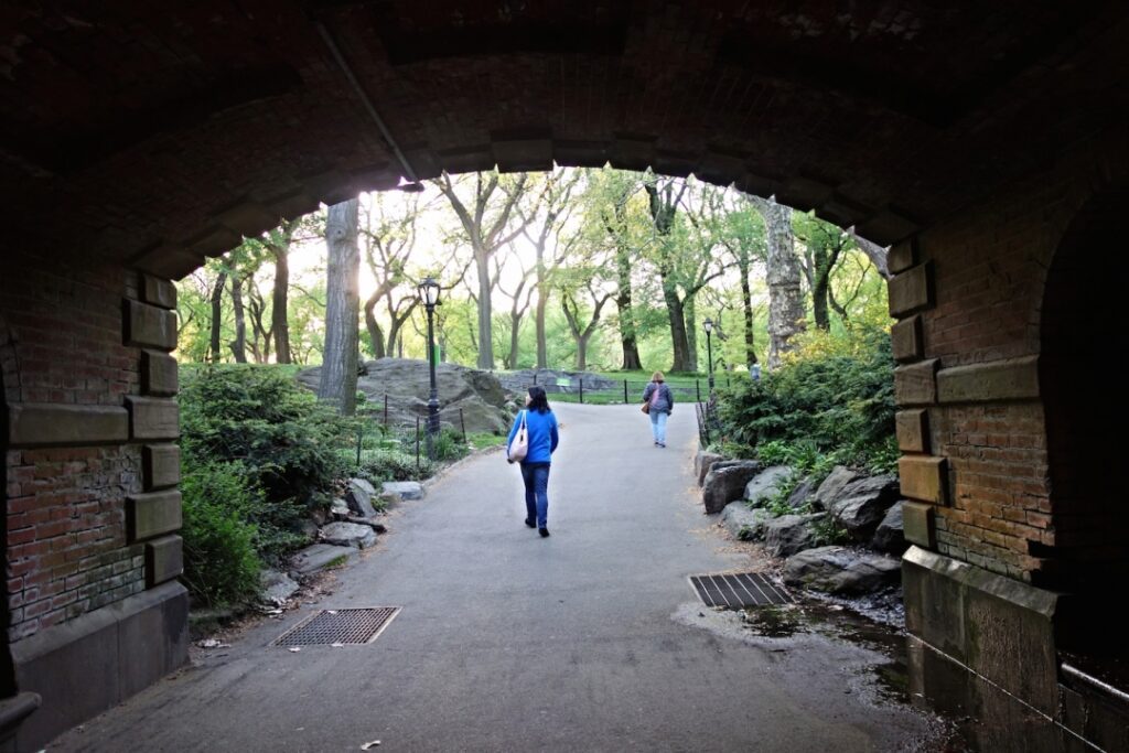 Two people walk on a paved path through a park, seen from under a brick archway, surrounded by trees, rocks, and greenery in daylight.