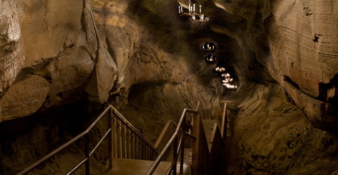 A metal staircase descends into a dimly lit cave with rough stone walls and several chandeliers hanging from the ceiling above.