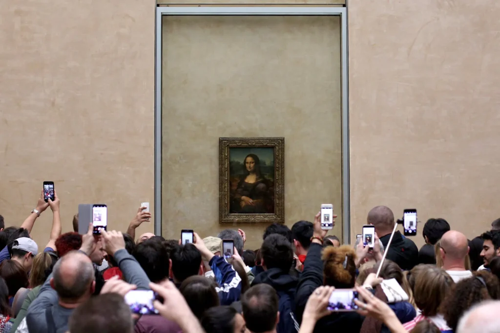 A crowd of people in a museum take photos of the Mona Lisa painting, which is displayed in a glass case on a beige wall.