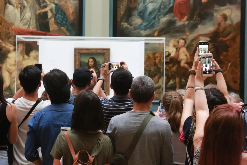 A crowd of people stands in front of the Mona Lisa at a museum, taking photos with their smartphones and cameras. The famous painting is displayed in a glass case against a backdrop of large classical artworks.