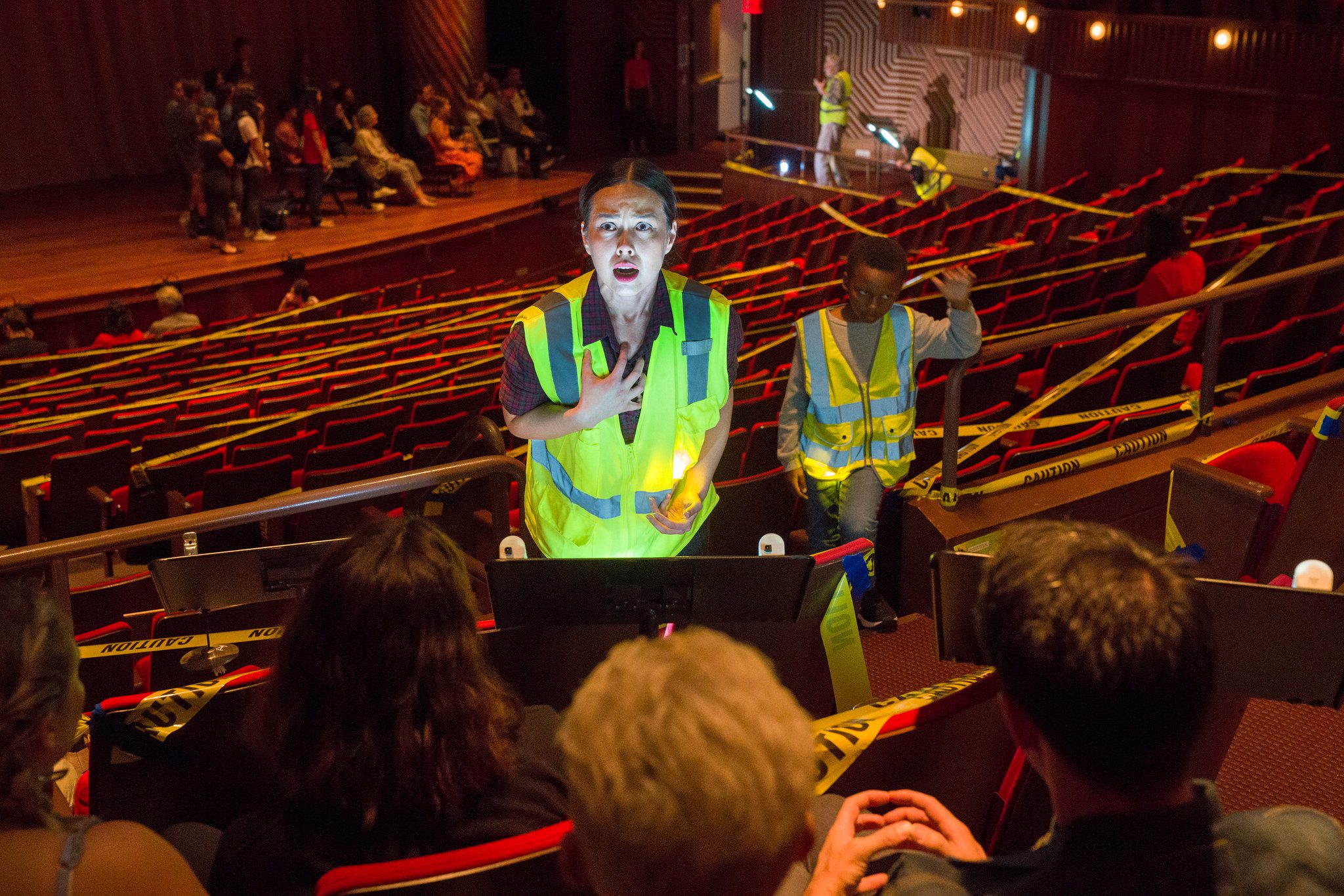 A woman in a reflective vest speaks to seated people in a theater with caution tape on rows. Another person in a vest stands nearby. The stage in the background has people gathered. The lighting is dim and dramatic.