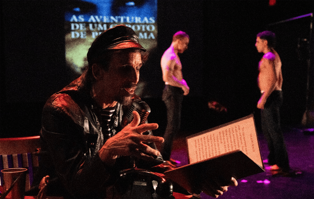 A person in a leather jacket reads from a book into a microphone onstage, while two shirtless people stand in the background with a projected image behind them.