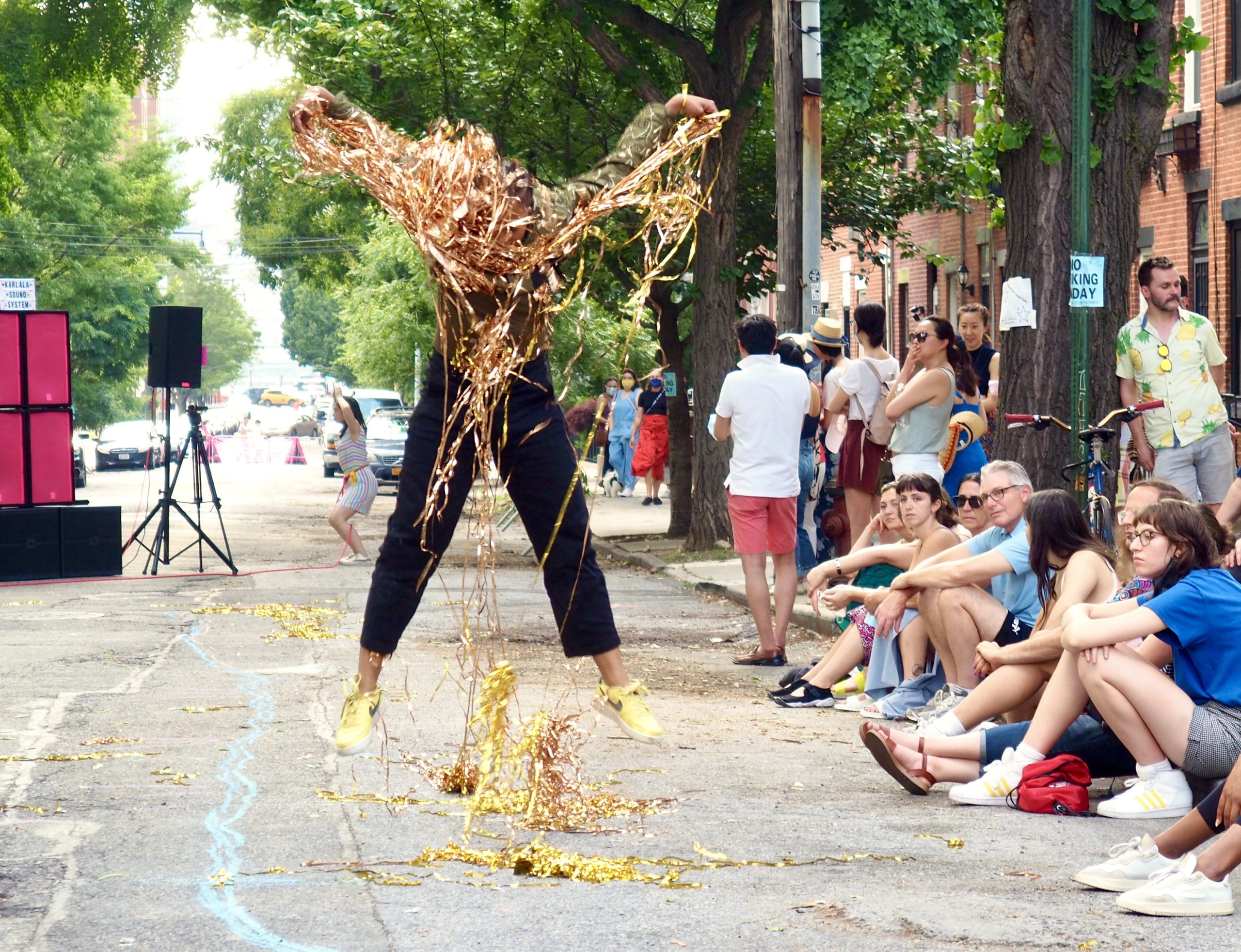 A performer jumping with their arms raised, in a cloud of gold tinsel. In the background, a group of people watching on a sidewalk.