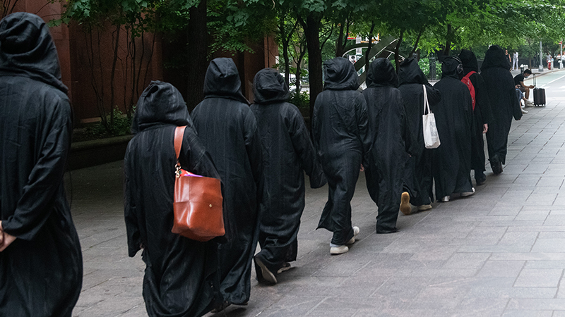 A line of people in black cloaks with their hoods up walking single file on NYU's campus.