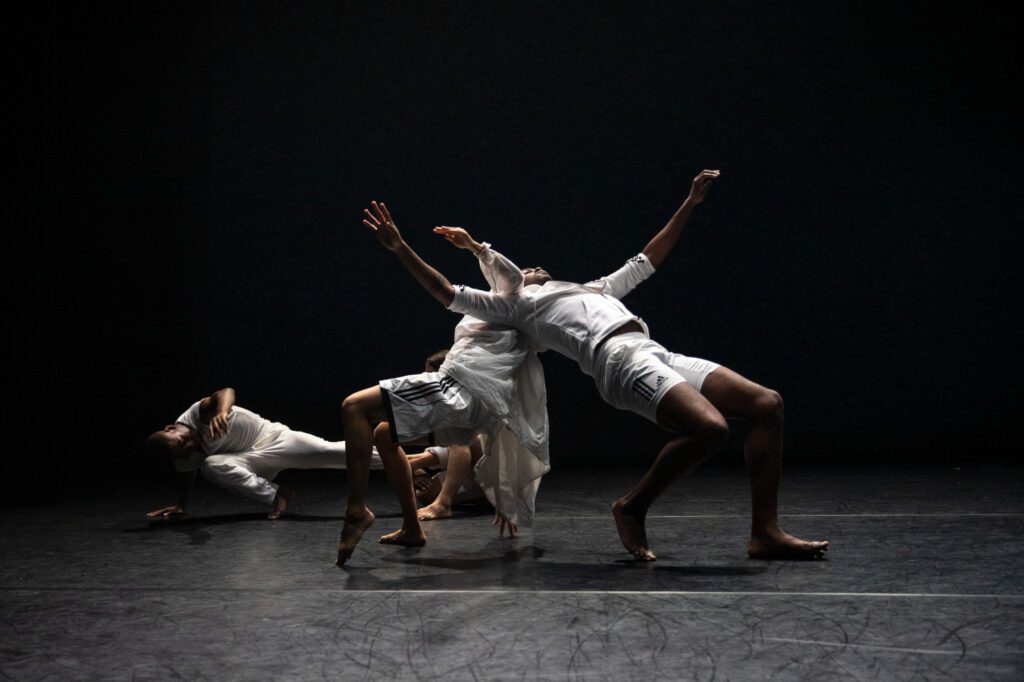 Four dancers in white perform on a dark stage. Two lean backward dramatically, one is bent forward, and another is lying on the floor, creating dynamic, expressive poses.