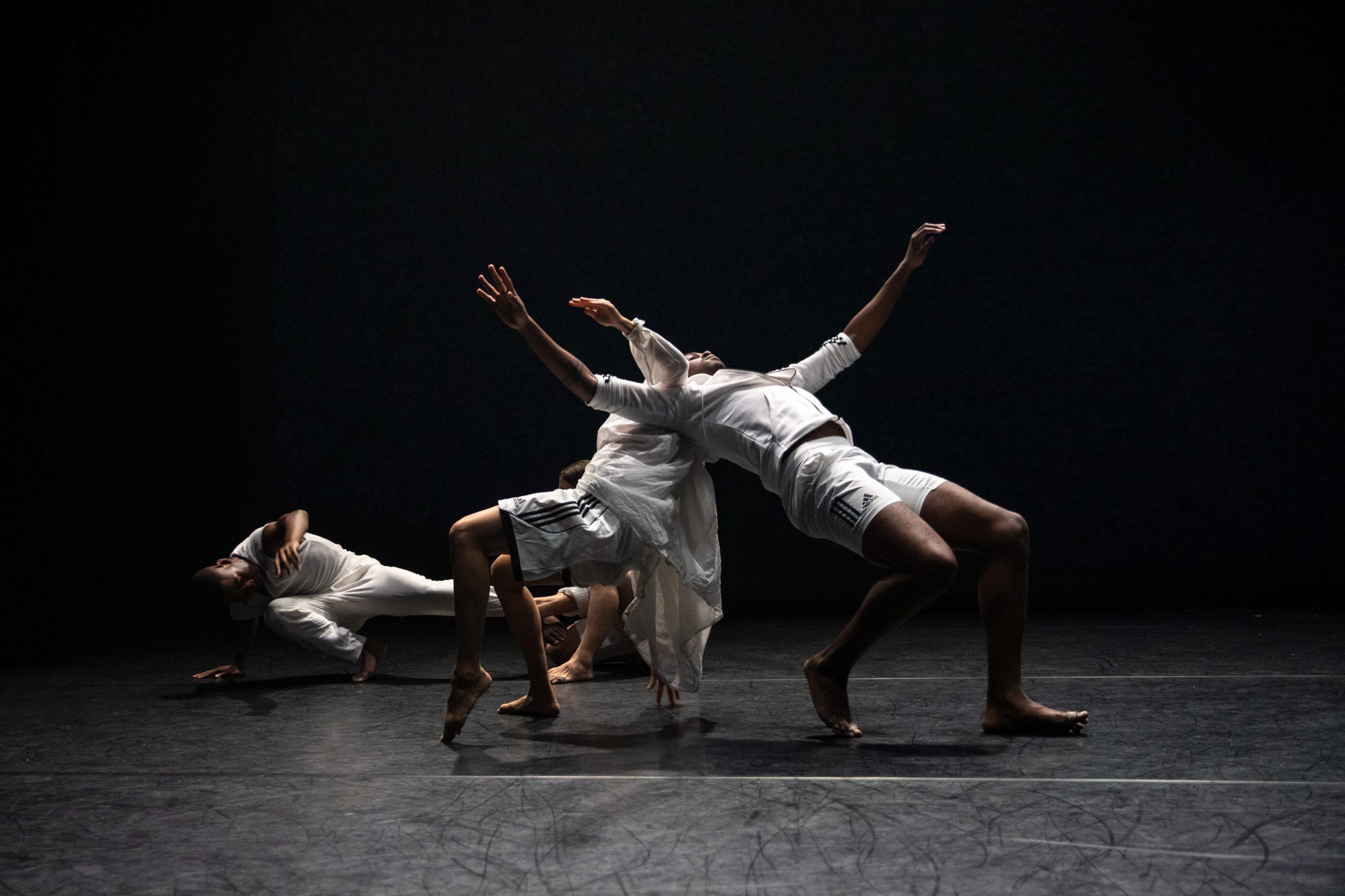 Four dancers in white perform on a dark stage. Two lean backward dramatically, one is bent forward, and another is lying on the floor, creating dynamic, expressive poses.