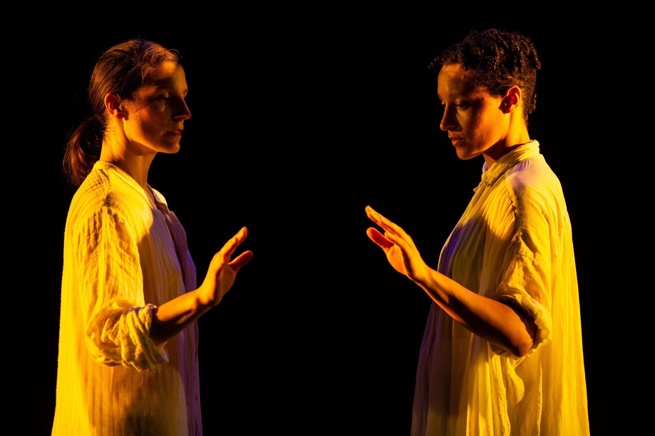 Two people in white shirts stand facing each other in dramatic lighting, each extending one hand forward as if about to touch, against a dark background.