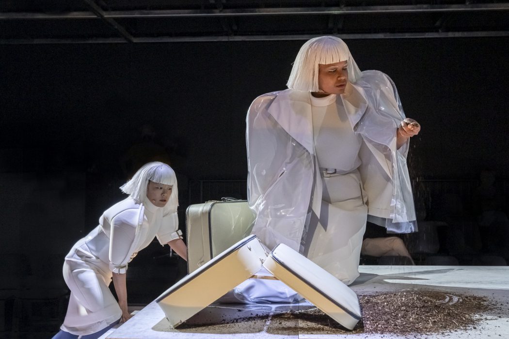 Two people in white wigs and futuristic white outfits interact with a suitcase spilling dirt onstage. One kneels with a suitcase, while the other stands with a handful of soil. The scene is set in a dimly lit, dramatic theater.