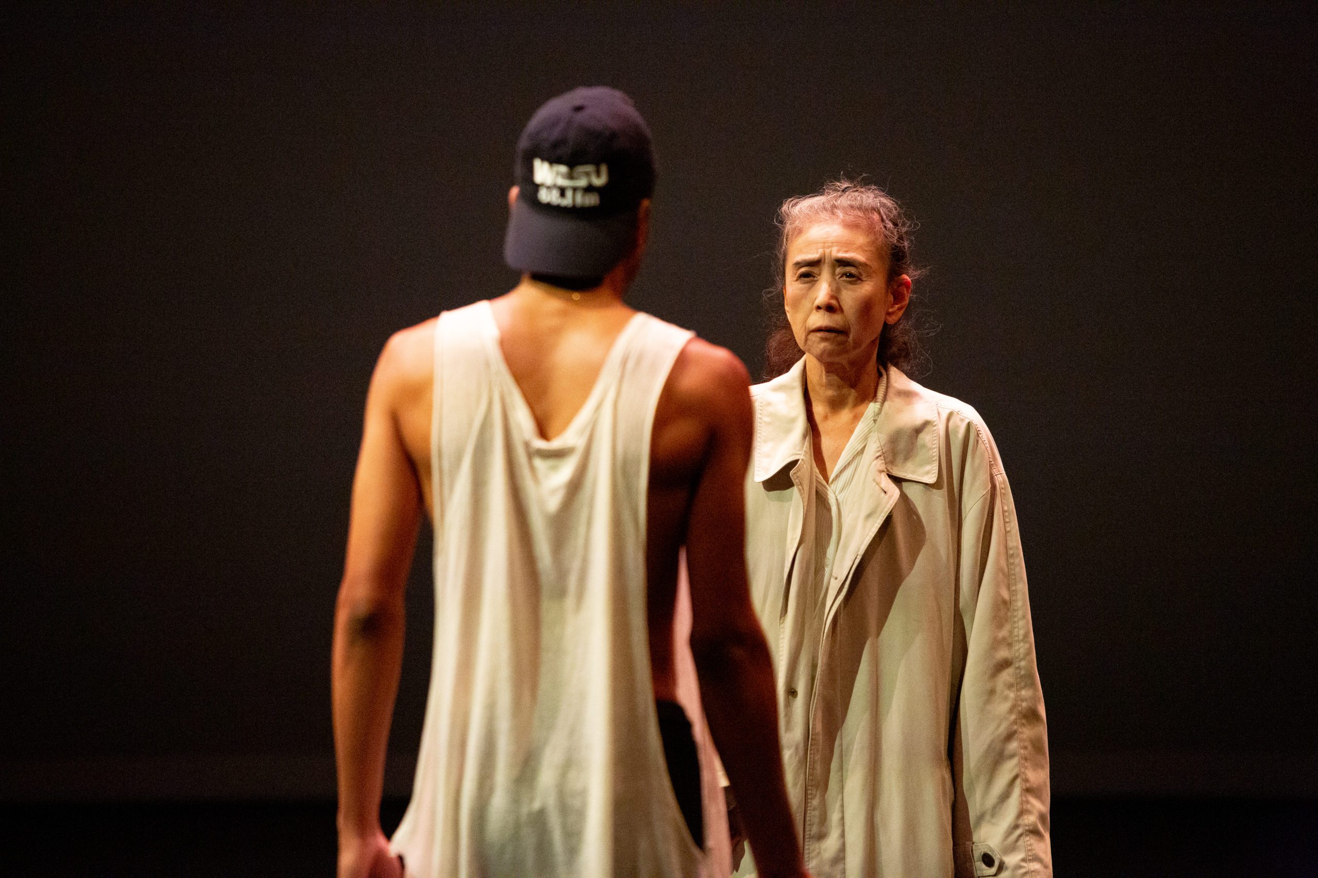 An older person in a beige coat looks concerned while facing a person in a sleeveless shirt and dark cap, standing under dramatic stage lighting.