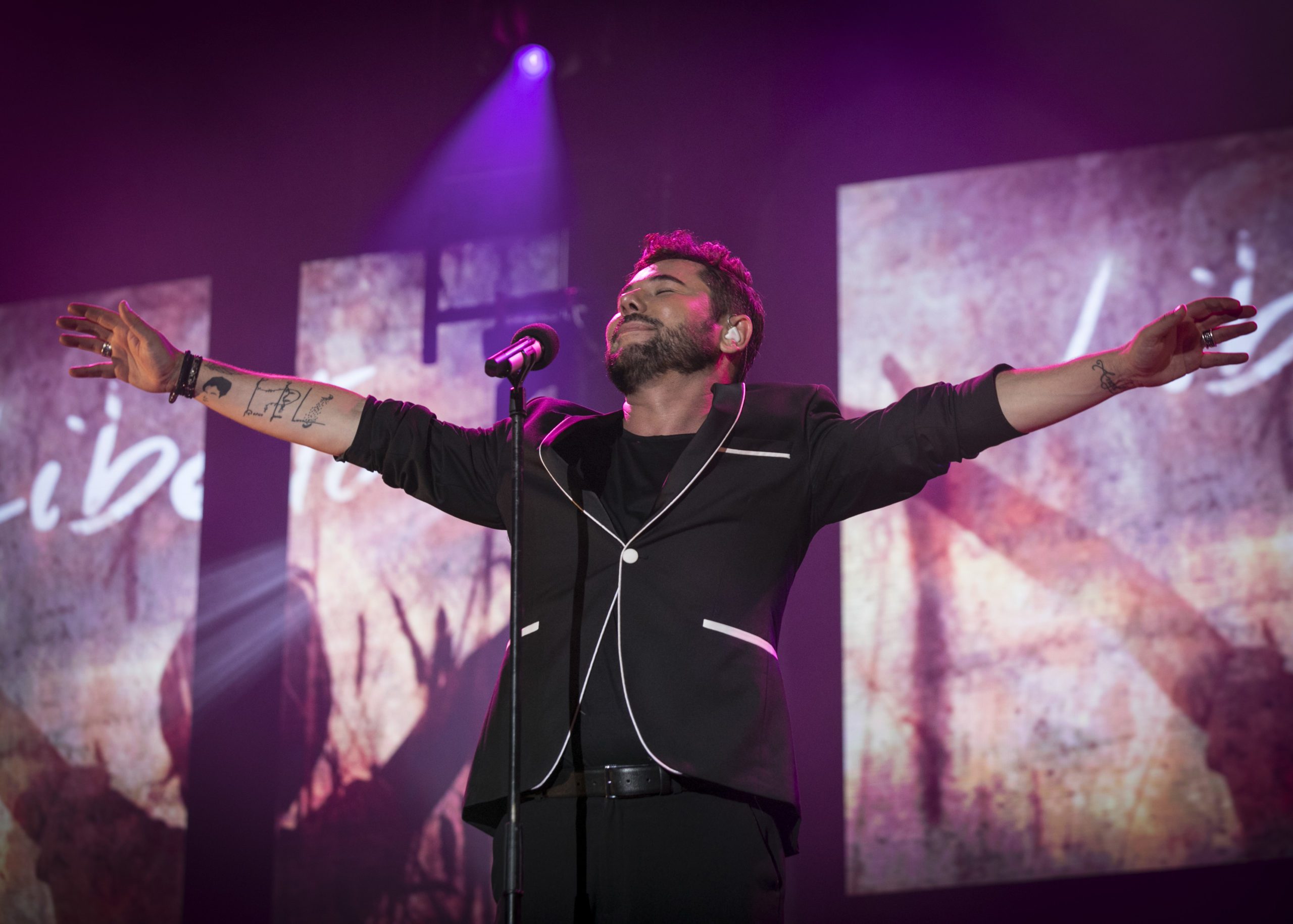 A singer in a black suit stands on stage with arms outstretched, eyes closed, and a microphone in front of him. Pink and purple stage lights and abstract screens are in the background.