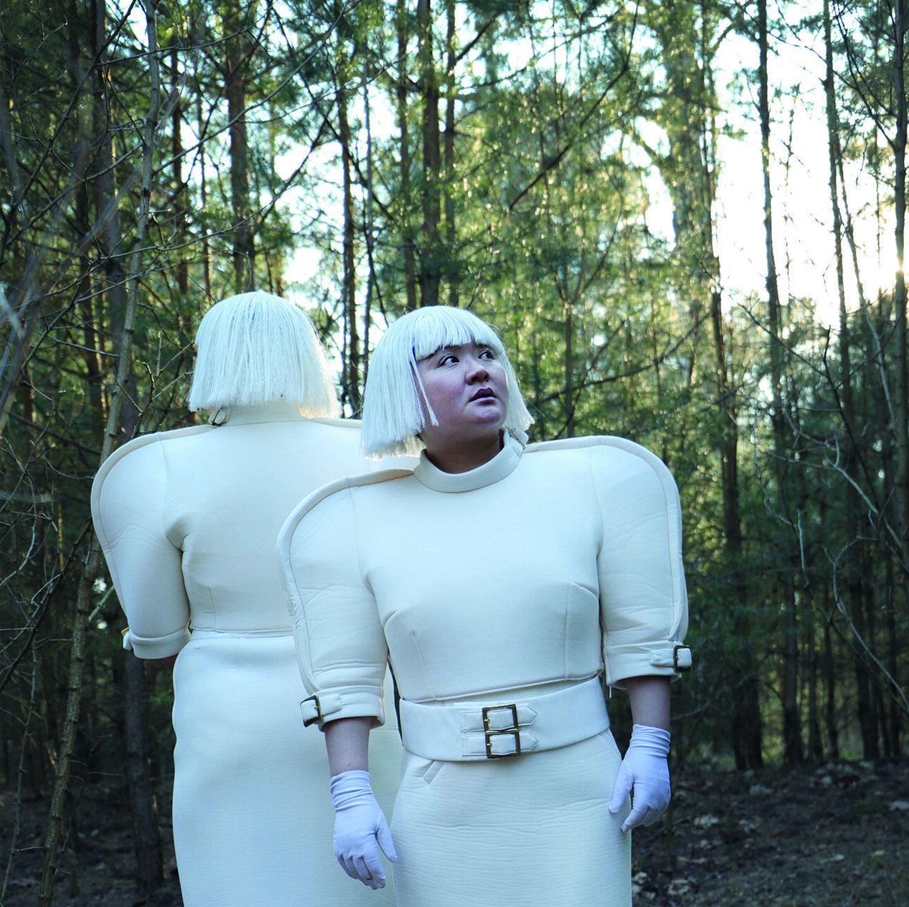 Two people with white bob wigs and futuristic white outfits stand in a forest. One faces forward, and the other faces away. Both wear blue tights, white gloves, and white shoes with chunky blue details.