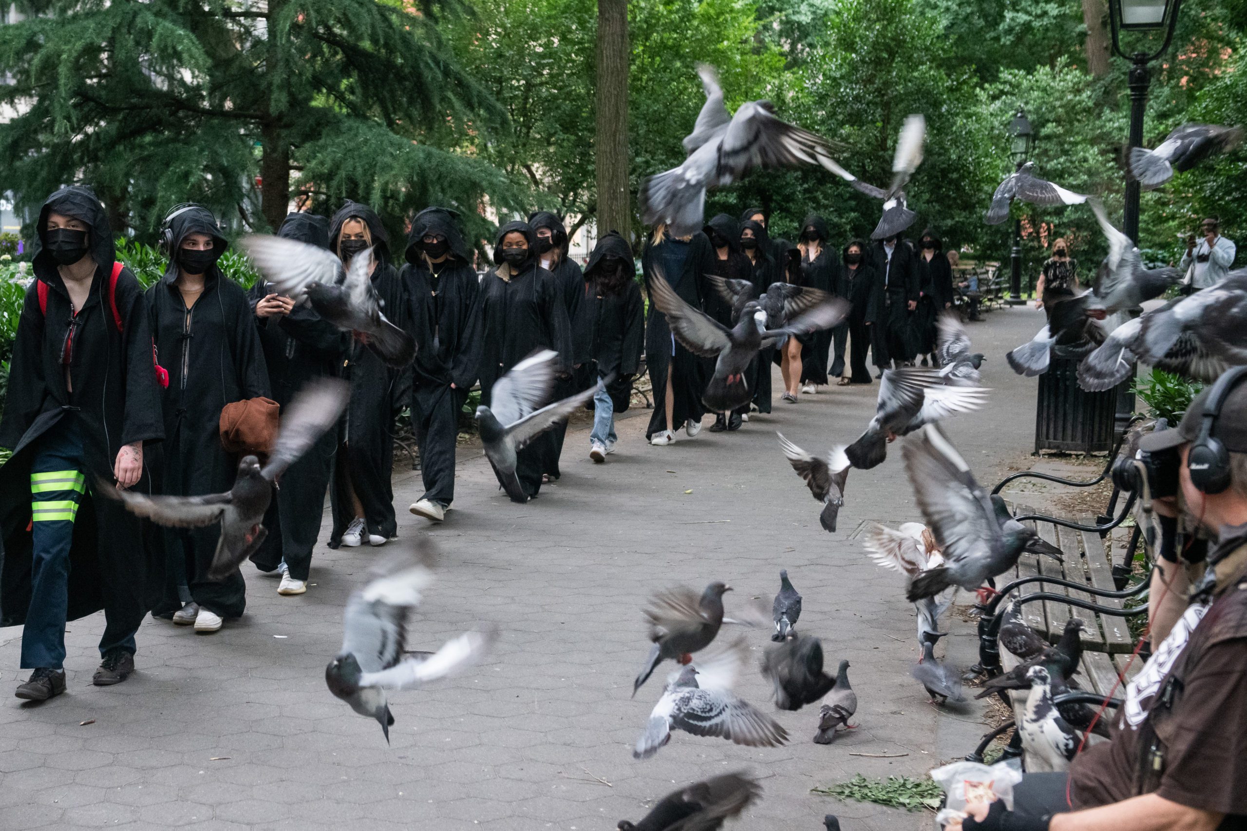 A group of people wearing black robes and masks walk through a park as pigeons fly up in front of them. Benches, trees, and greenery surround the path. Some bystanders are seated, watching the scene.