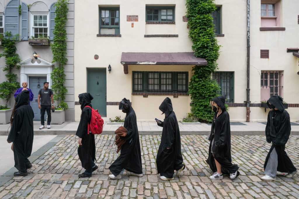 Six people in black hooded cloaks walk in a line on a cobblestone street. One carries a red backpack, and another looks at their phone. Two bystanders watch from a doorway in the background.