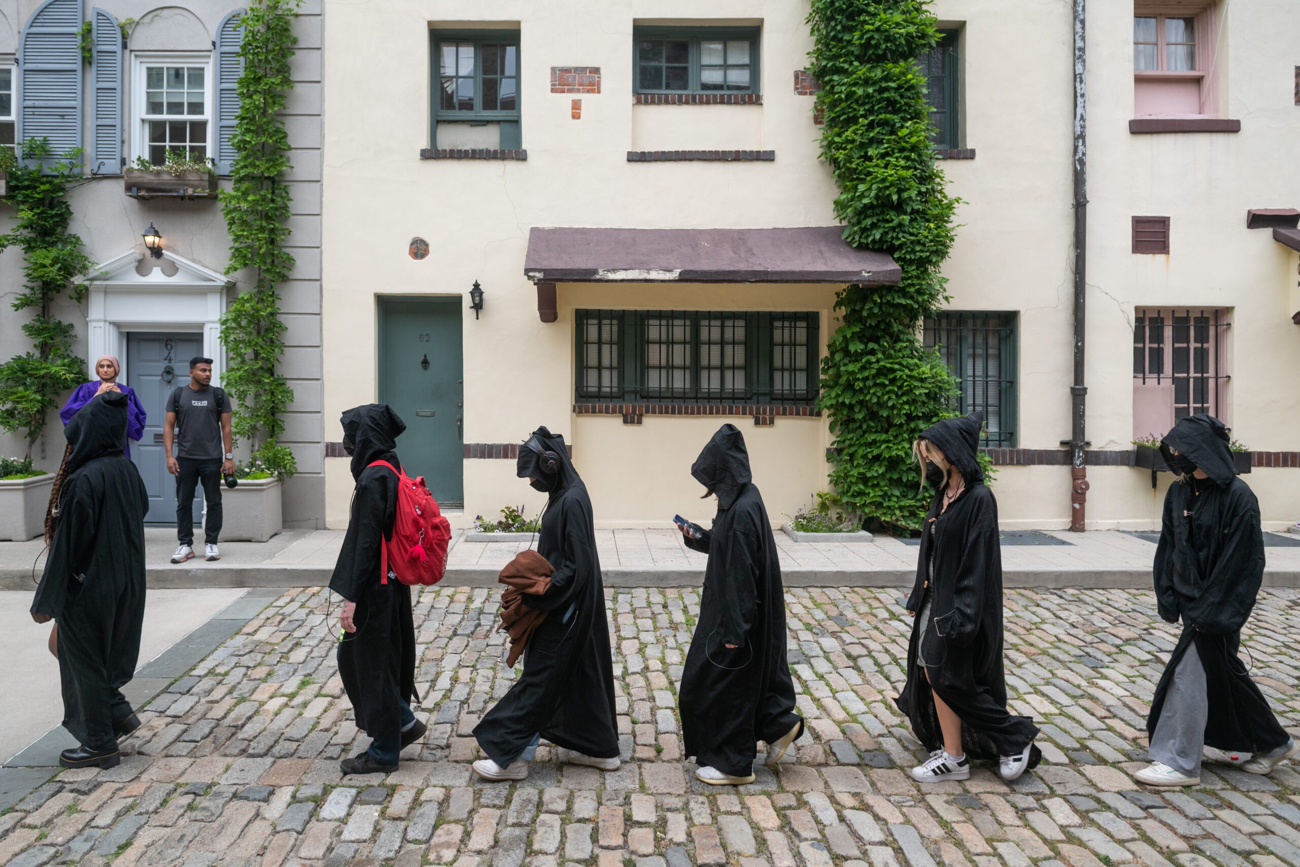 Six people in black hooded cloaks walk in a line on a cobblestone street. One carries a red backpack, and another looks at their phone. Two bystanders watch from a doorway in the background.