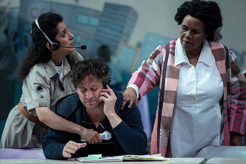 A man in a dark shirts sits at a table frowning while on the phone while two women stand behind him. One is hugging the man while looking at the other woman who is putting a hand on the man's shoulder with a face of distress.