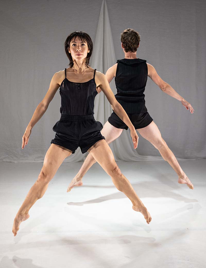Two dancers wearing black, with rigid limbs. One facing the camera, one facing a draped curtain in the opposite direction.