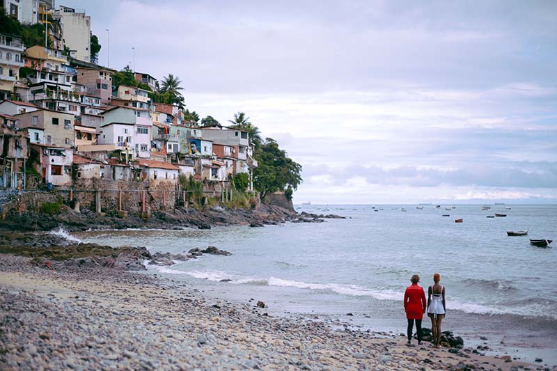 Two people stand with their back facing the camera at a sand shore. The water extends as far as the eye can see, and to the left homes are stacked atop one another an a rocky hill.