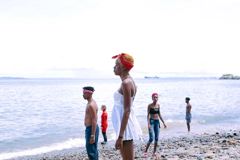 Five people stand on a sandy shore with the ocean extending in the background. The woman closest to the camera is wearing a white dress and an orange bandana, a bit back and behind her is a man with no shirt wearing a red bandana, another woman wears a red long dress and another wearing jeans with a black bandeau top, at the far right background stands another shirtless man..