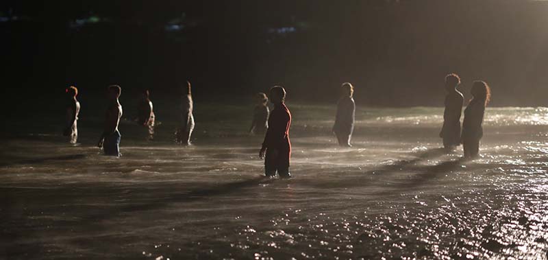 A group of people stand in a circular position in the water at the shore. They face each other and the lighting is dark.