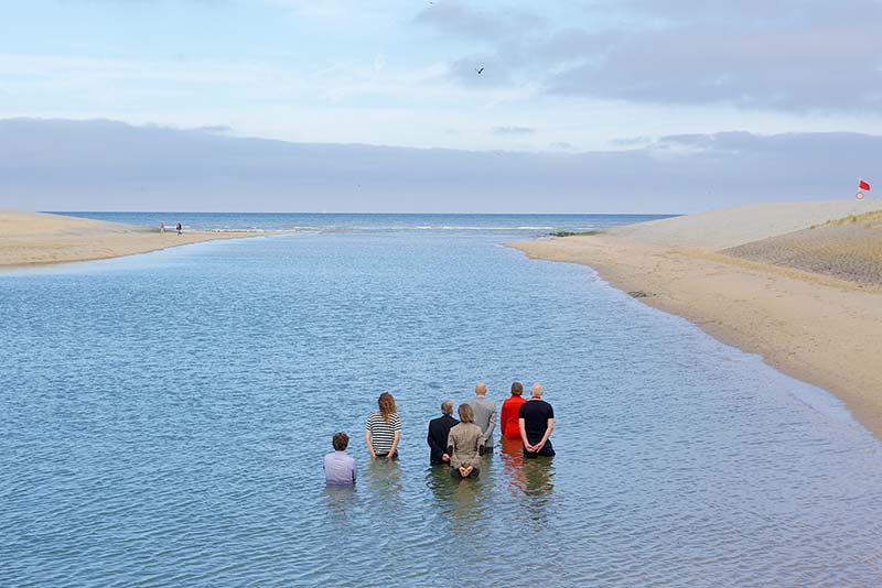 A group of 7 people standing in the sea with their backs facing the camera. Water is about waist high. Sand is visible a few feet away at the shore.