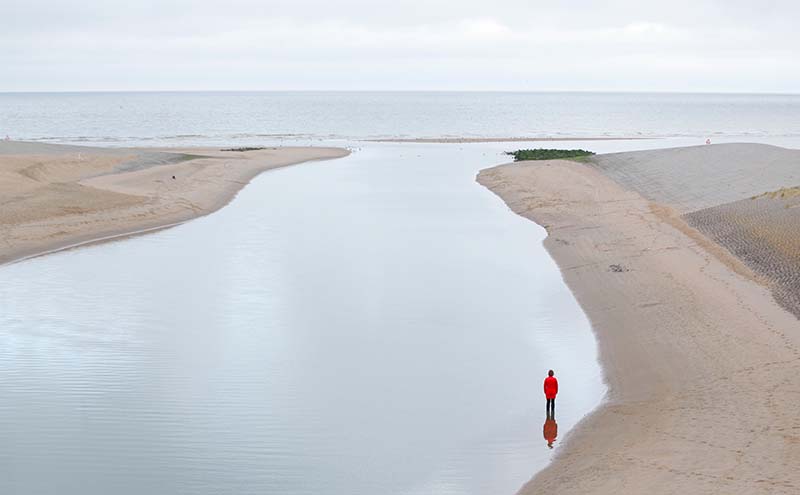 A water shore with someone in red standing at the edge of the sand.