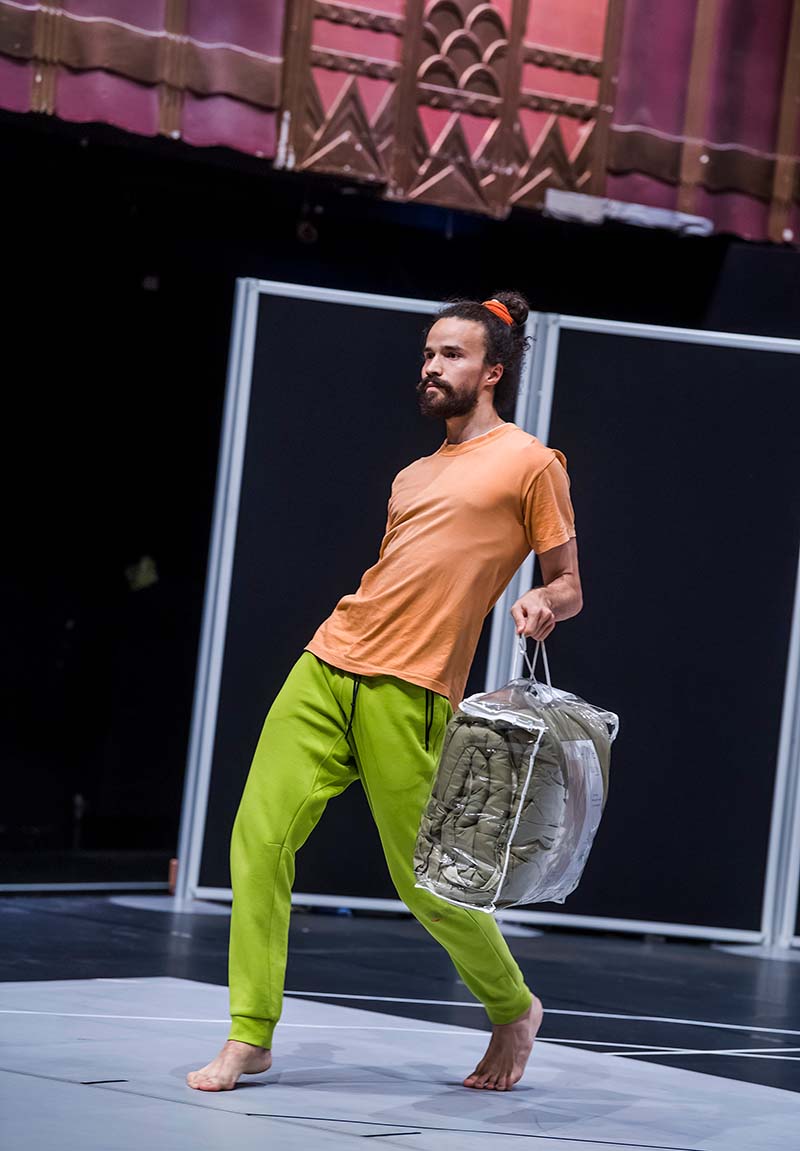 Man with green pants and orange tshirt holding a box with one hand in a slanted dance position.