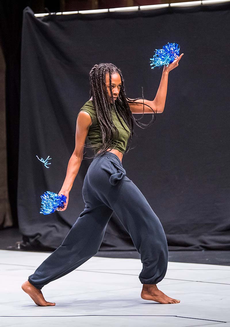 A woman with pants and a green shirt dancing wiith blue small fans in each hand.