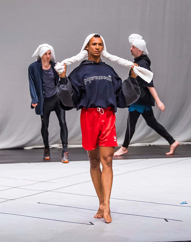 A person wearing bright red shorts, a dark blue jacket, and a white towel on their head walks down a runway with two more people who are dressed similarly but with pants are behind in line to follow.