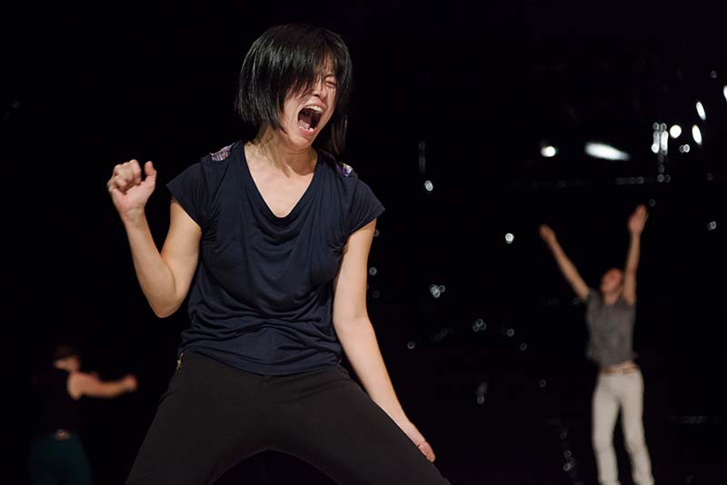 A dancer in a black shirt stands at the center of the picture screaming with someone in the background whose arms are raised towards the sky.