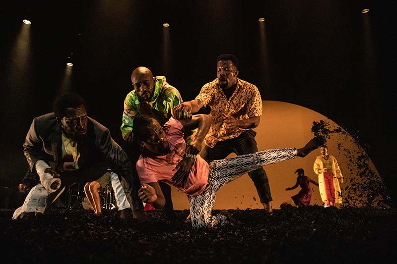 A group of dancers wearing colorful clothing stand at the edge of the stage with one lower and raising his leg while the others hold him.