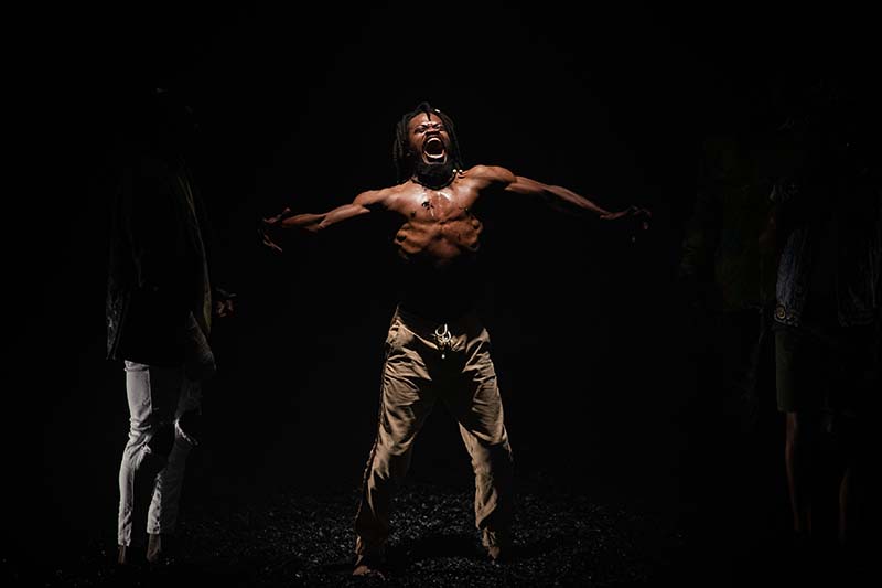 A shirtless man standing in the middle of a black background with his arm extended to the side.