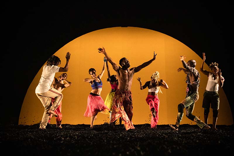 A group of dancers in colorful clothing stand in a circle with arms raised in middle of dance. The background is dark with a yellow centered half circle.