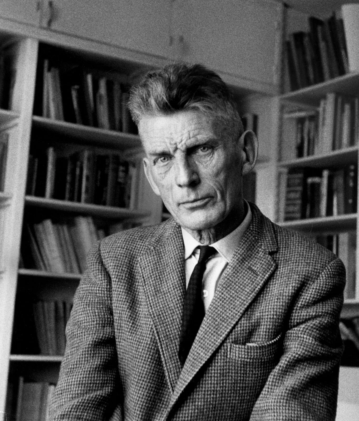 A man with short hair and a serious expression sits in front of bookshelves filled with books, wearing a checkered suit, white shirt, and dark tie. The image is in black and white.