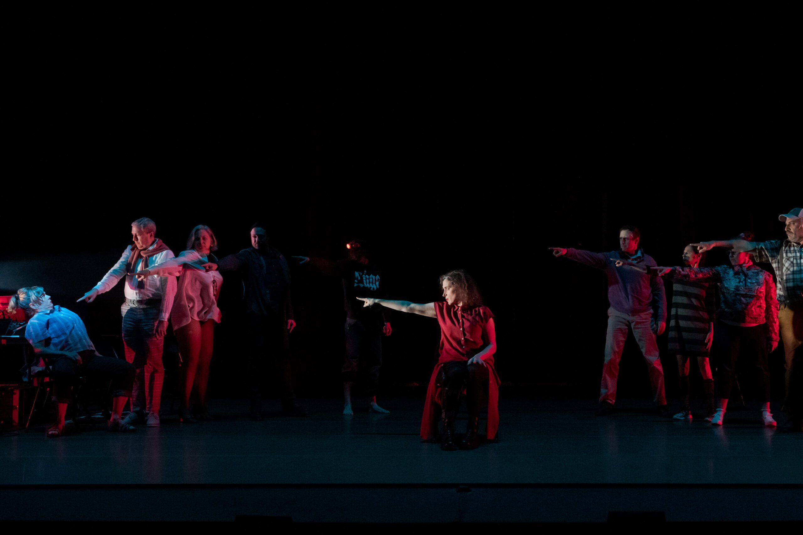 A group of people on stage, mostly standing and pointing outward, with one person in a wheelchair in front. The scene is dramatically lit with red and blue lights against a dark background.