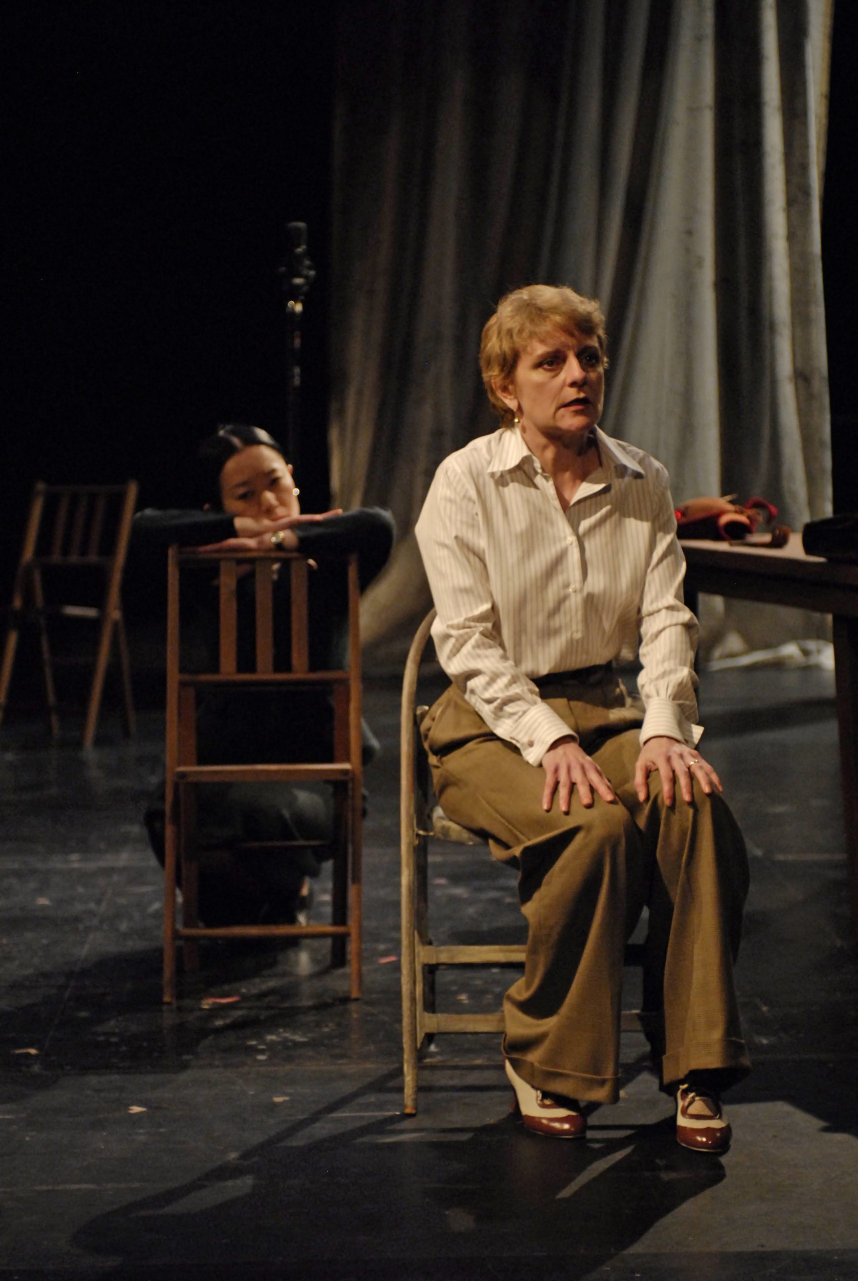 A woman in tan pants and a white button down sitting in a chair facing the camera. To her left is a person sitting backwards in a chair admiring her.