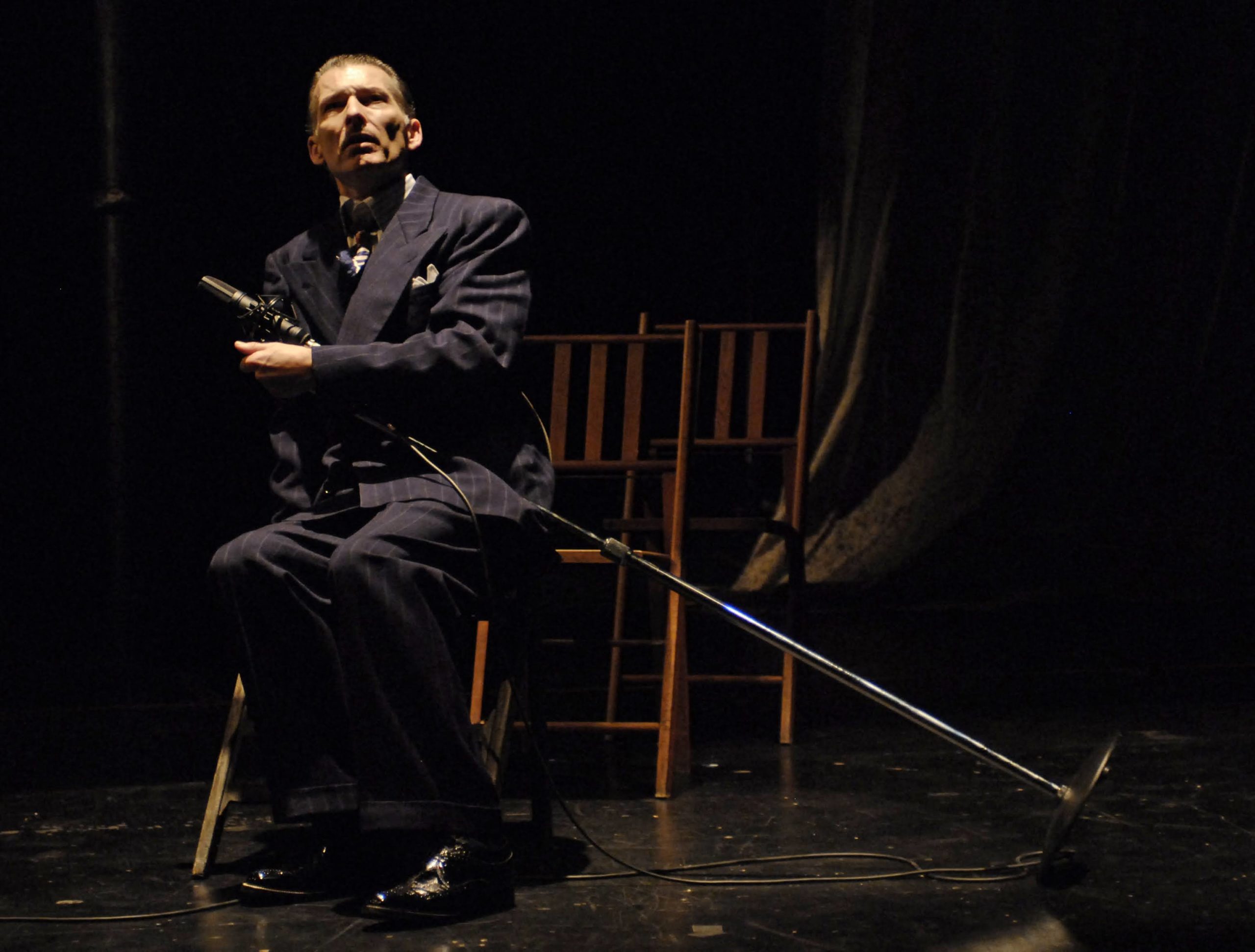 A man to the left of the frame wearing a full dark suit sitting in a chair holding a microphone stand that is leaning on his lap.