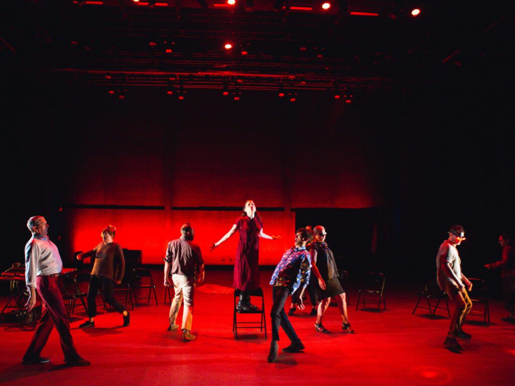 Seven people perform onstage under dramatic red lighting; one person stands on a chair in the center while others pose around them, some leaning back, creating a dynamic, theatrical scene.