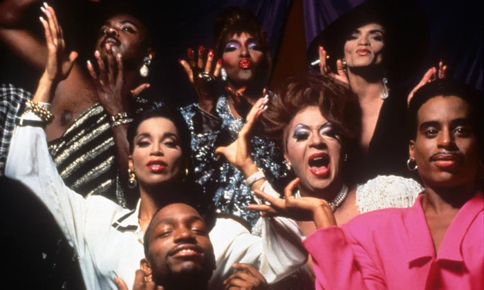 A group of people wearing glamorous outfits and makeup pose dramatically, striking expressive and celebratory poses against a dark background.